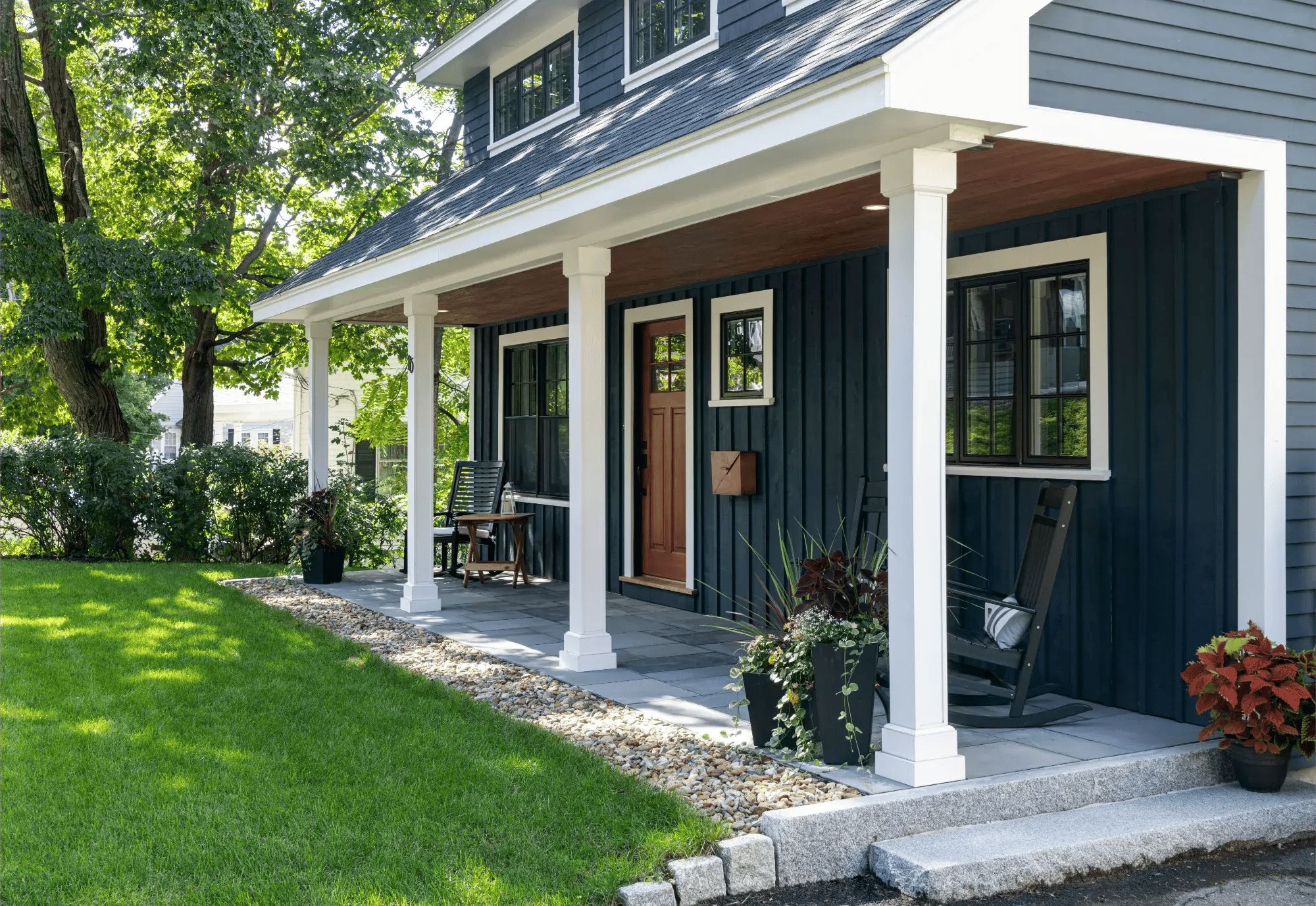 Front porch of a custom home in Greater Boston with dark blue siding, white columns, wood ceiling, and landscaped entryway